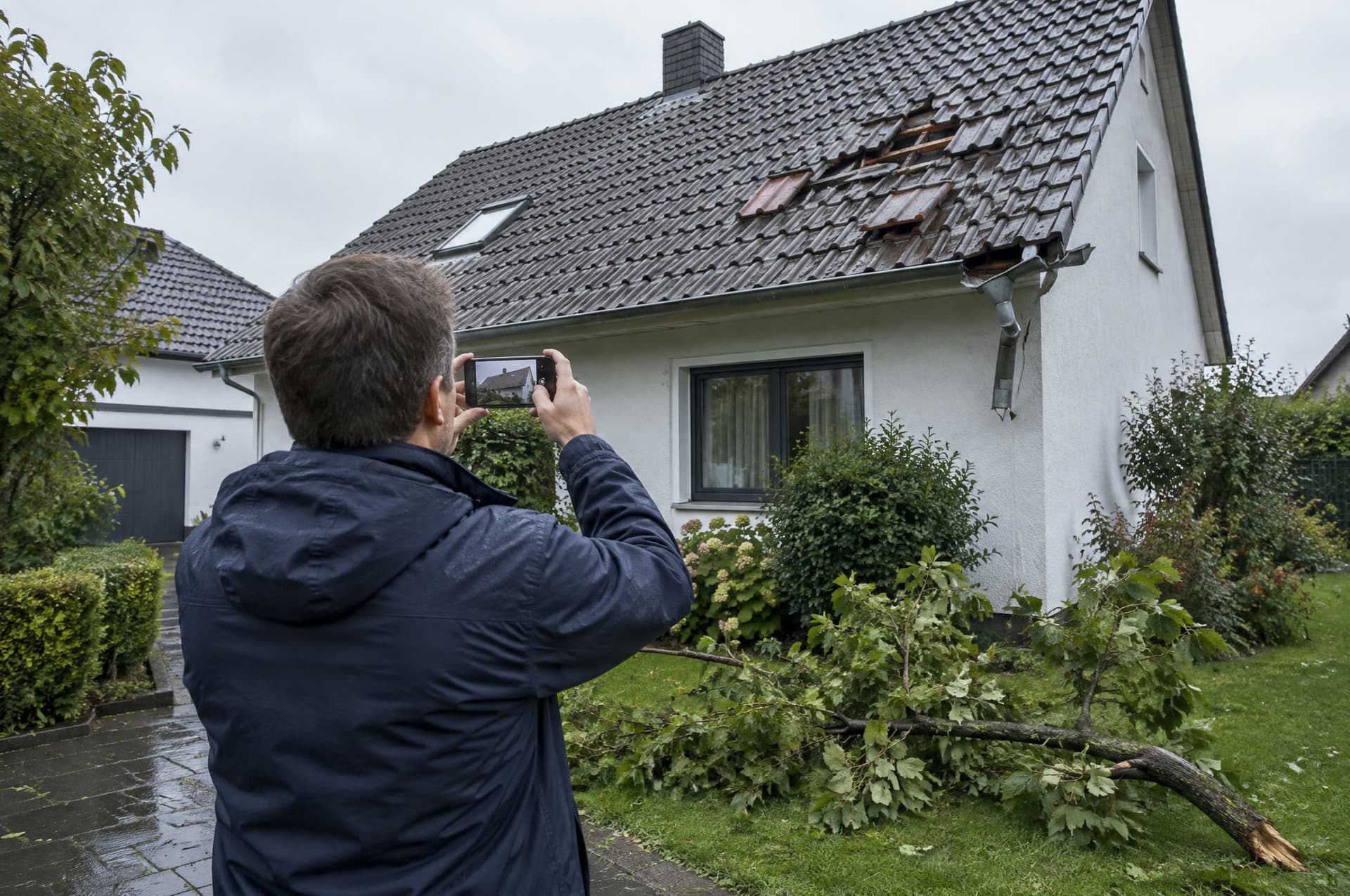 Ein Schadenregulierer von hinten fotografiert mit dem Smartphone einen Sturmschaden am Dach eines Einfamilienhauses, im Garten liegt ein abgebrochener Ast.