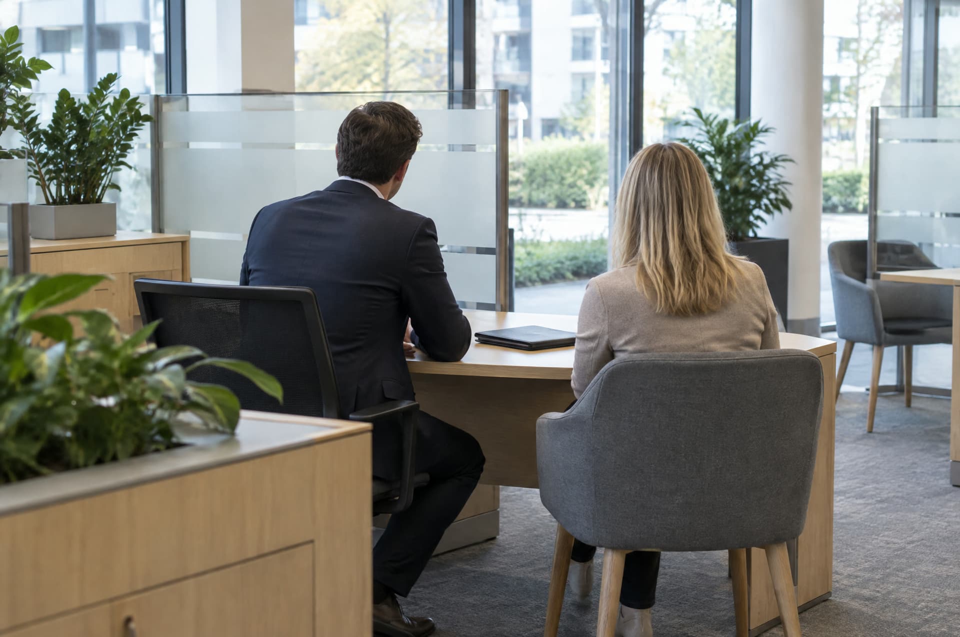 Consultation area of a bank branch with light wooden desks and glass partitions, an advisor and a customer seen from behind talking at a desk.
