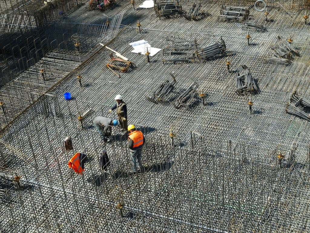 Blick von oben auf eine Baustelle: Bewehrungsstahl einer Bodenplatte, mehrere Arbeiter mit Helmen und Warnwesten.