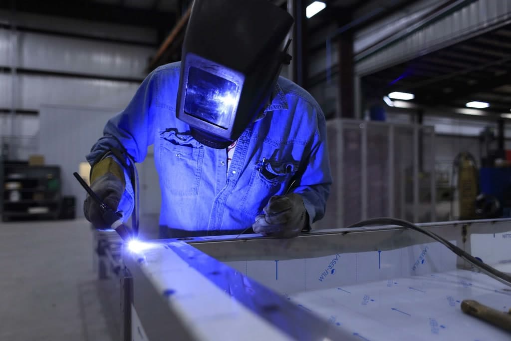 A welder in a helmet and blue workwear welding a sheet-metal part in a workshop hall.