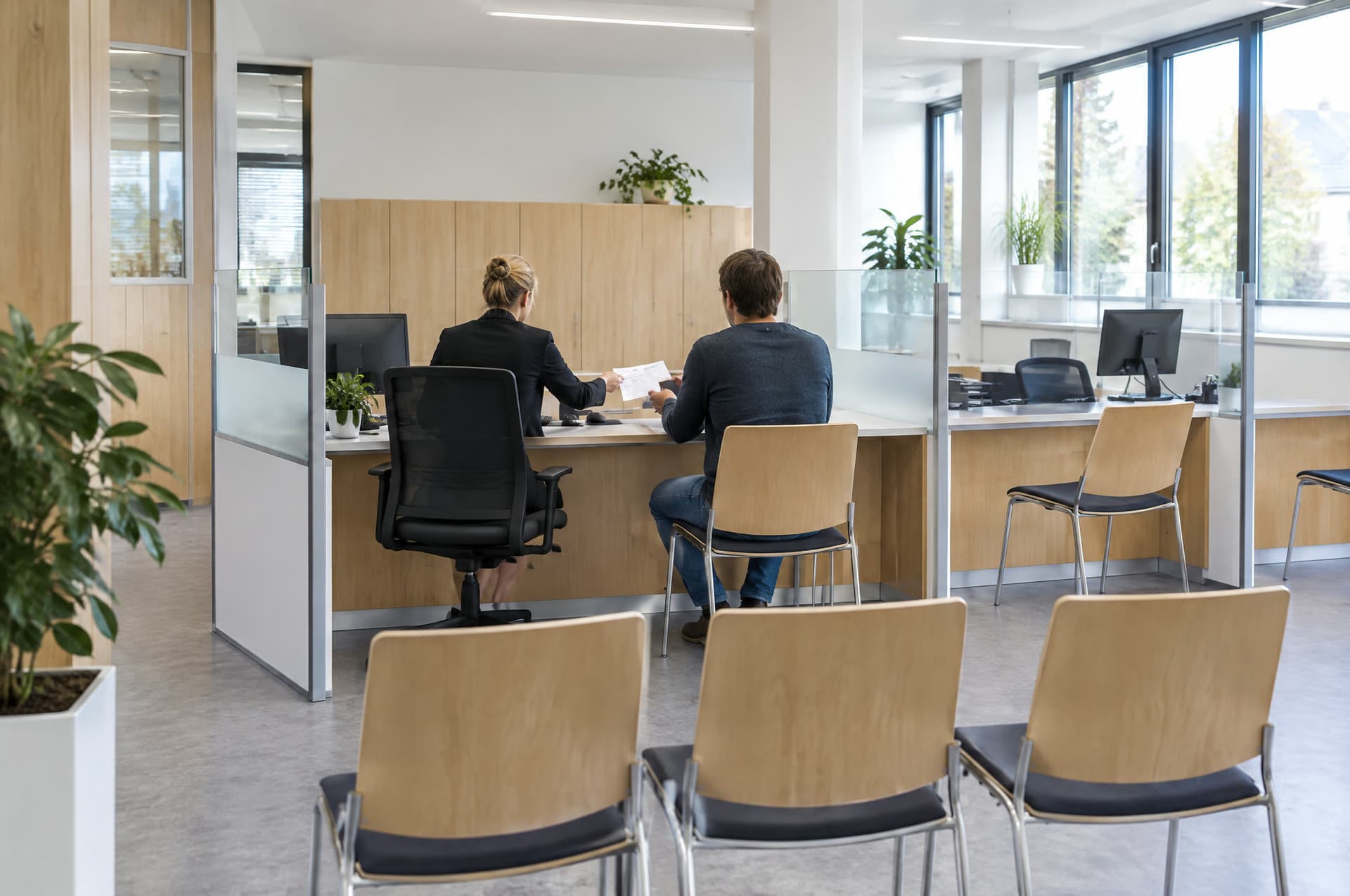 Bürgerbüro mit Beratungsplätzen und Wartebereich, zwei Personen von hinten an einem Schreibtisch, eine reicht ein Formular.