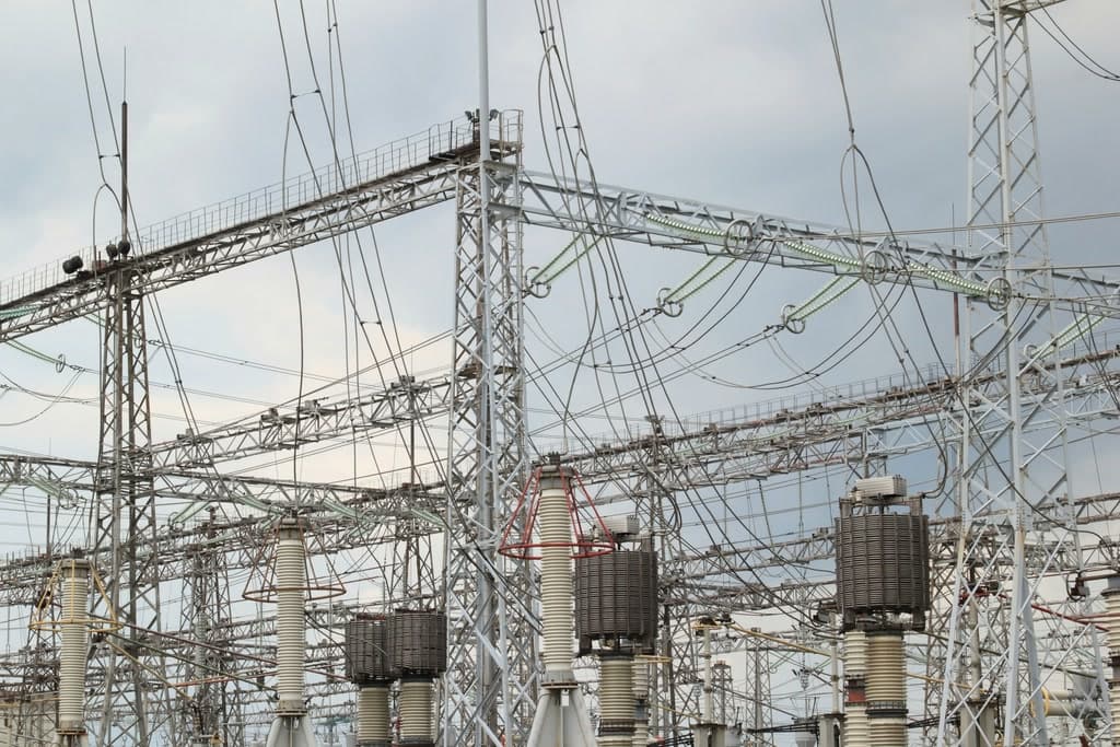 Electrical substation with lattice masts, lines, insulators and transformers against a cloudy sky.